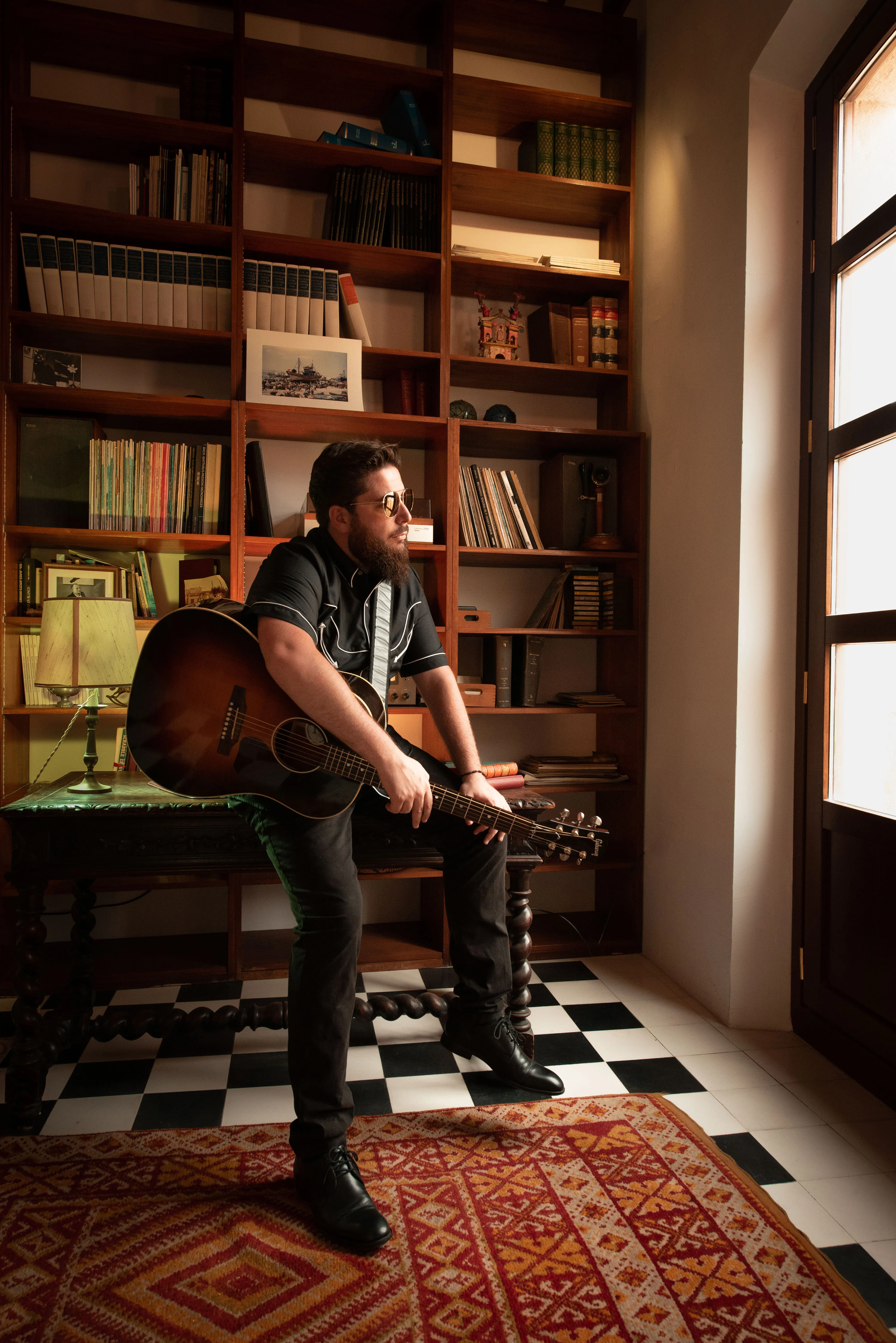 JJ Fuentes sentado con una guitarra acústica ante una estantería de libros, en una casa con suelo de damero